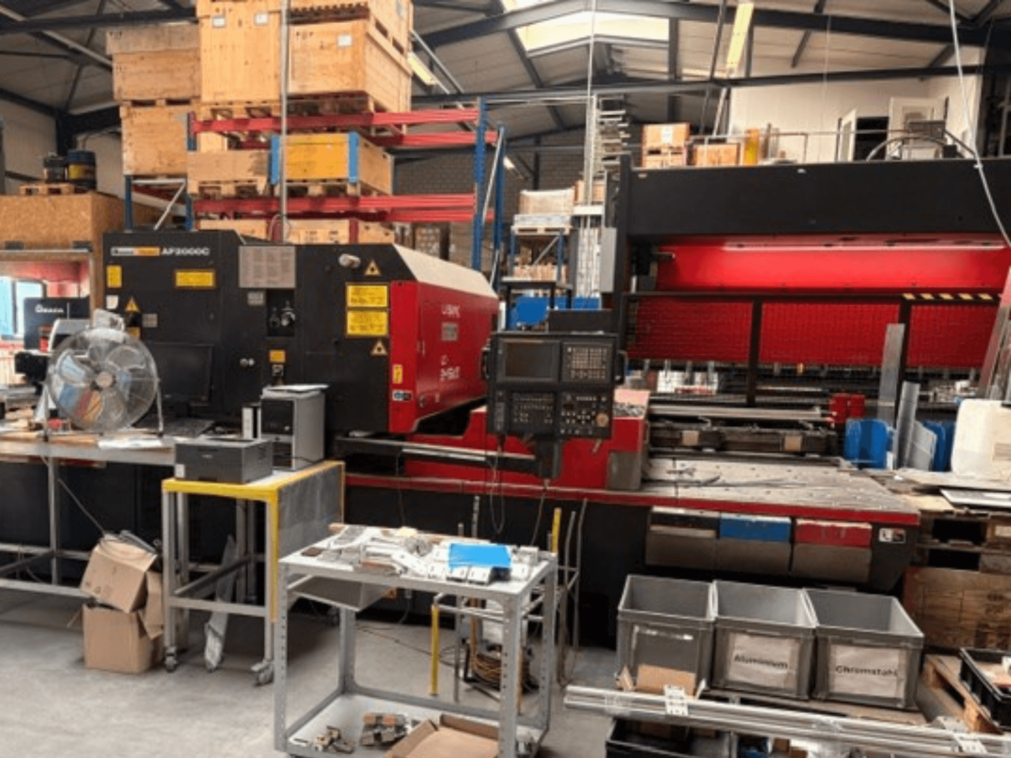AMADA LC-2415αIII CO₂-lézervágógép műhelyi környezetben, elölnézetben, a vezérlőpanellel és a munkaterülettel együtt.
