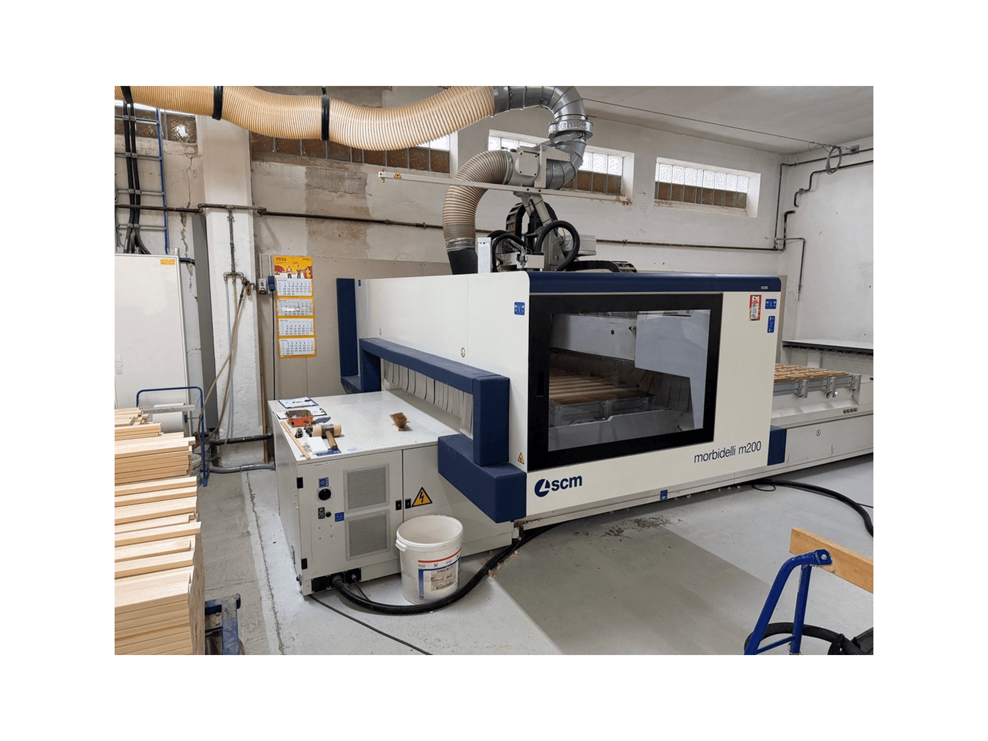 SCM Morbidelli M200F CNC megmunkáló központ egy műhelyben, oldalnézetben, az asztalon fadarabokkal és a felette elhelyezkedő szellőzőrendszerrel.