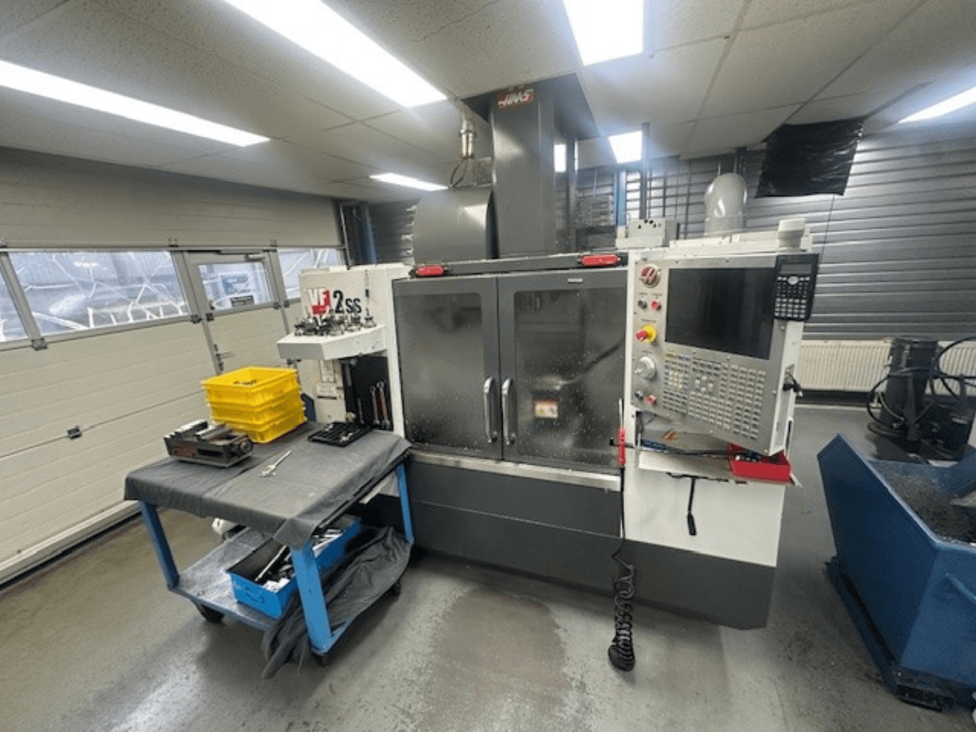 A HAAS VF-2SS függőleges megmunkálóközpont elülső nézete a vezérlőpanellel, a munkaasztallal és a szerszámtároló állomással.