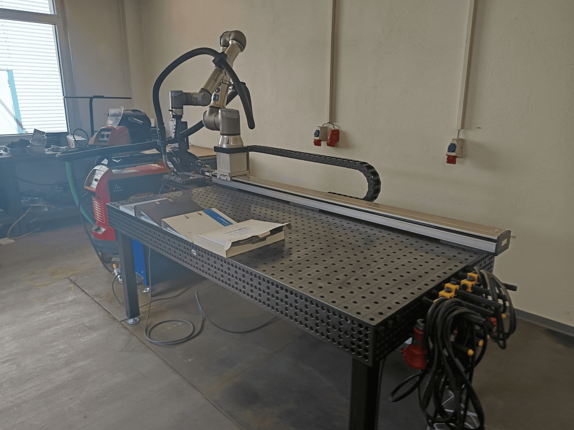 A FRONIUS TransSteel 4000 impulzushegesztő cellája oldalról nézve, robotkarral, munkaterülettel és áramcsatlakozásokkal.