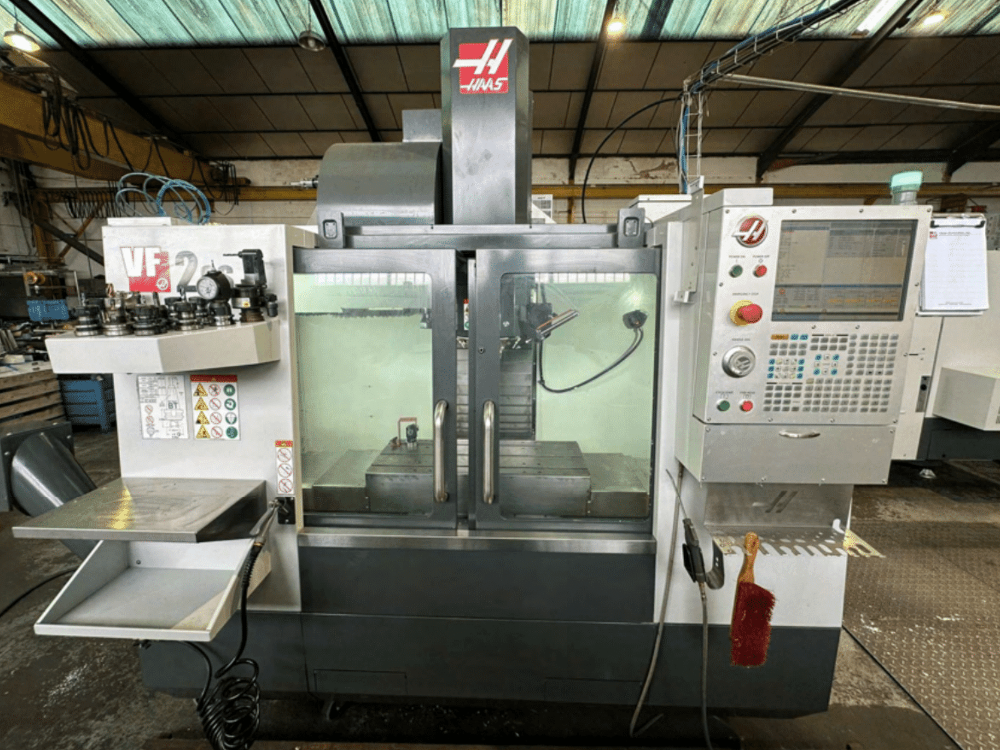 Haas VF2 CNC-gép előlnézetben, a vezérlőpanel, a szerszámtartók és a munkaterület biztonsági címkékkel és kefével.
