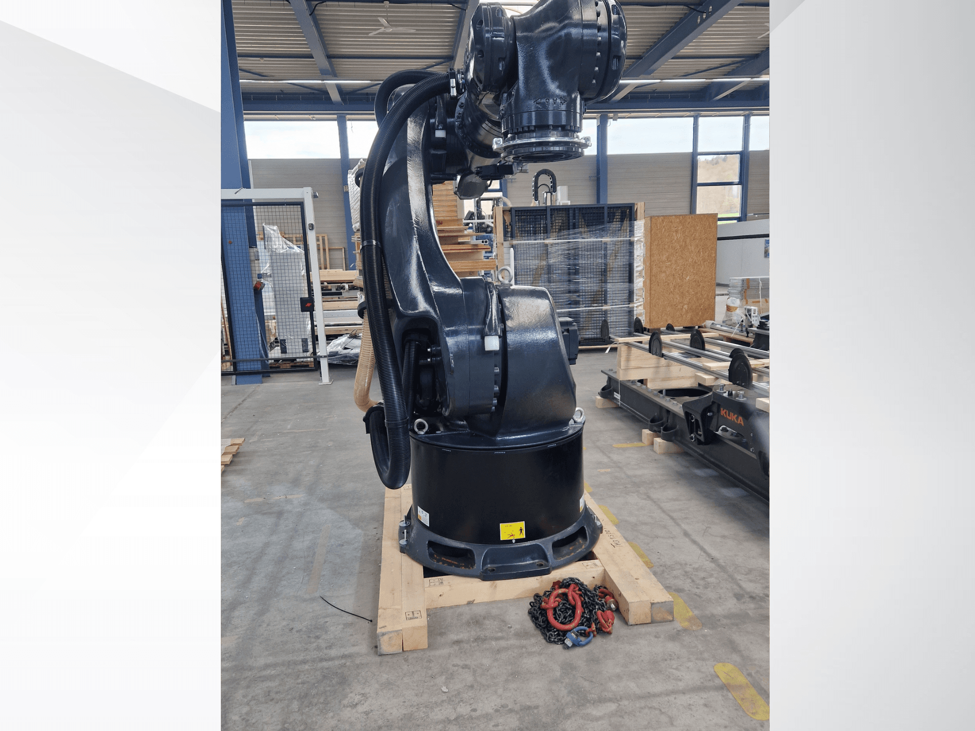 KUKA KR 340 R3330 robotkar egy raktárban, oldalnézetből, amelyen látható az alapja és a kábelek, az előtérben láncokkal.
