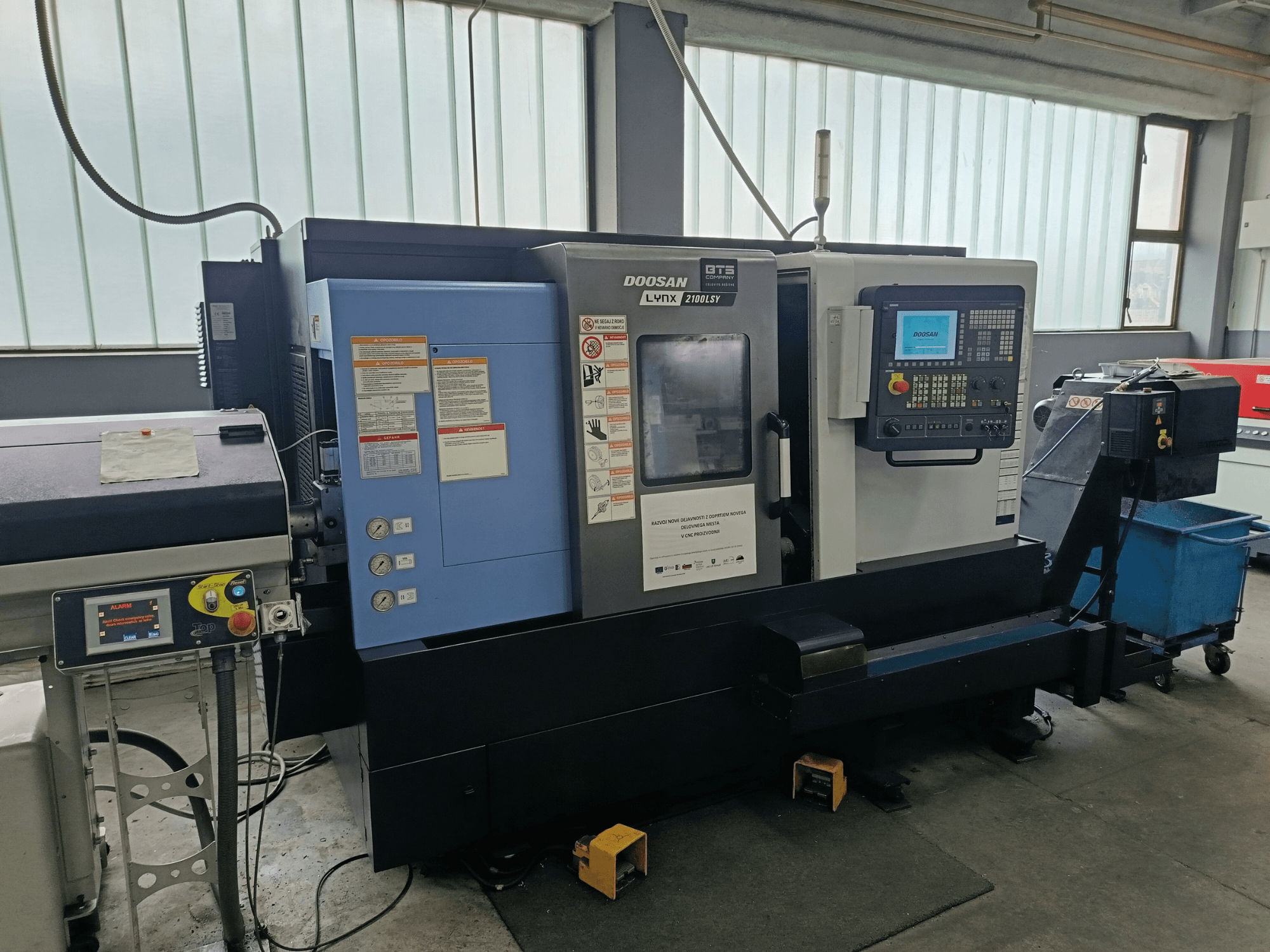 DOOSAN LYNX 2100 LSY esztergapad-központ, oldalnézet a vezérlőpanel és a kezelőgombok, látható információs címkékkel.