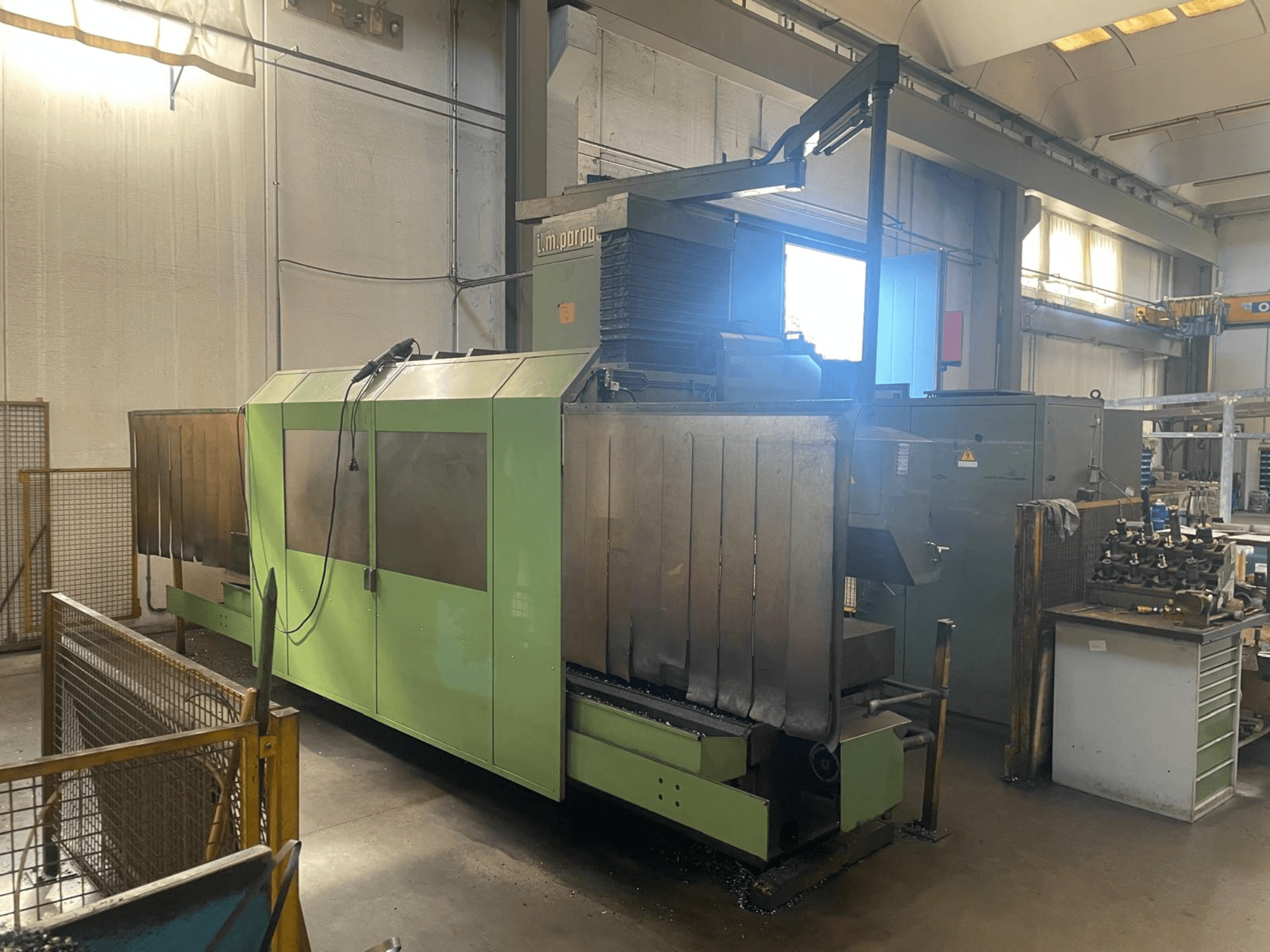 Zöld ipari gép látható fém alkatrészekkel, elölnézetben; vezérlőpanel, biztonsági figyelmeztetések és a közelben lévő munkaállomás.