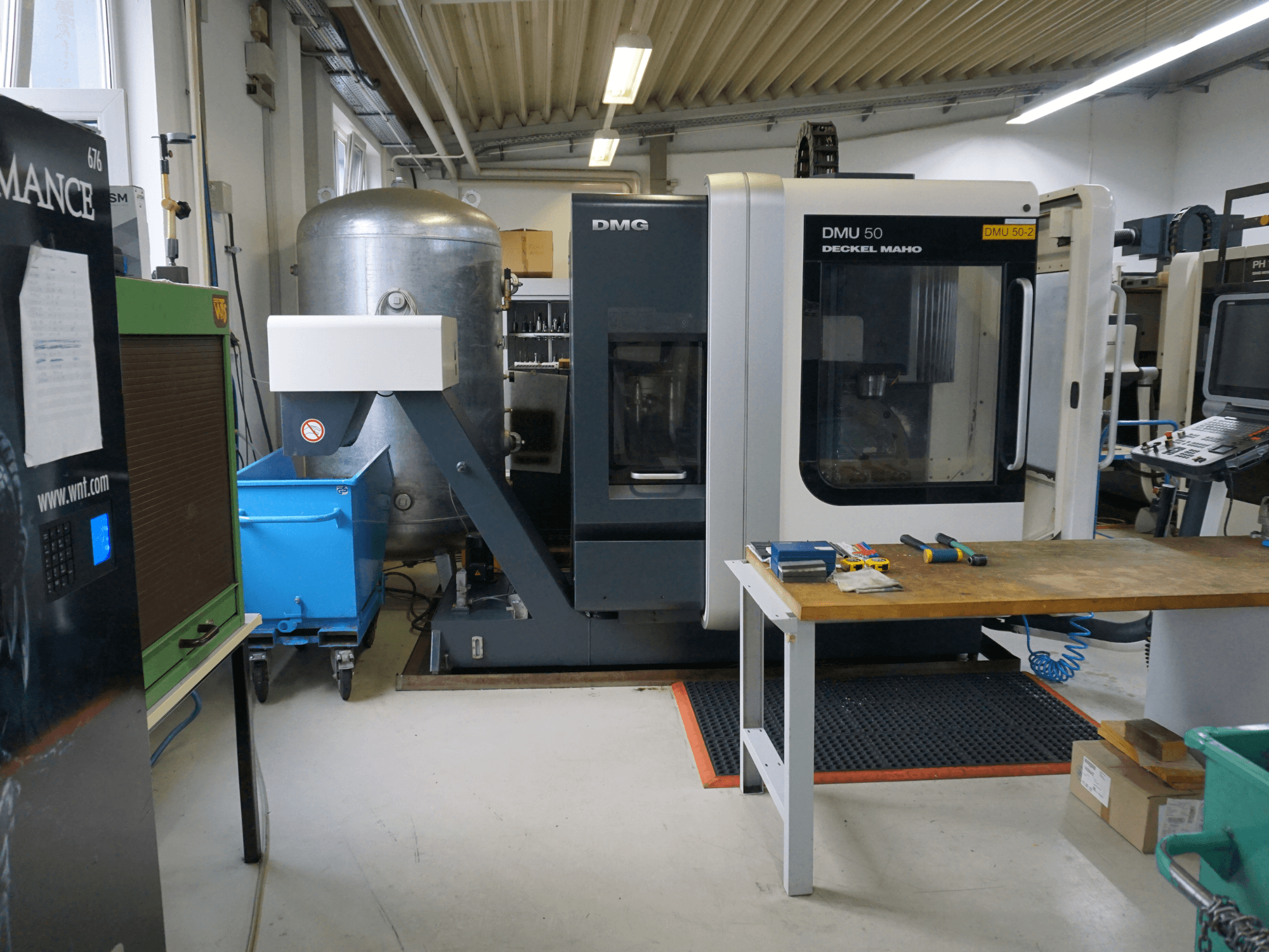 DMG DMU 50 marógép egy műhelyben, elölnézetben, szerszámokkal egy faasztalon és egy kék kerekes szemetesládával a közelben.