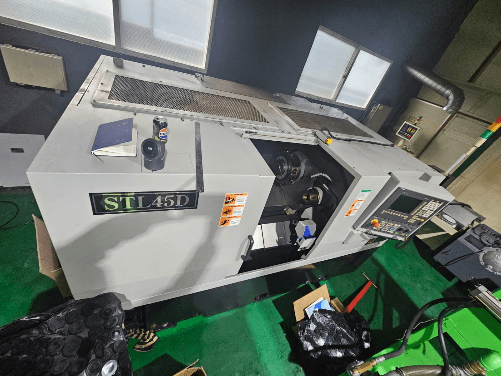 STL45D CNC-gép felülről nézve, a vezérlőpanel, a hengeres alkatrész, a notebook és a tetején lévő kanna látható.