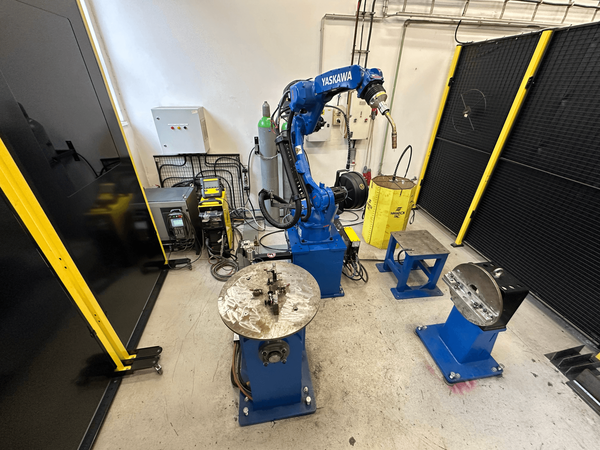 Yaskawa robotkar kék színben, két munkaasztallal, hegesztőberendezésekkel és biztonsági korlátokkal; ipari munkaterület elölnézetben.