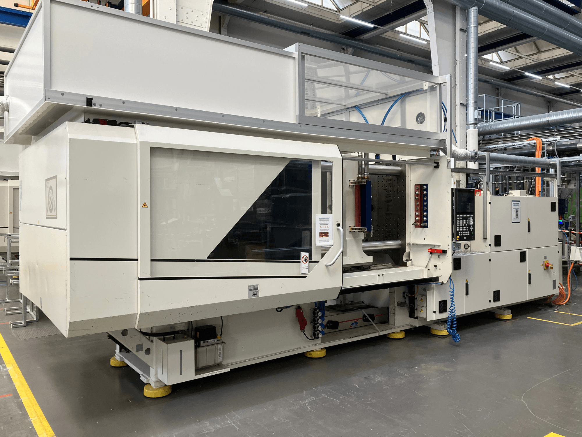 Egy AET típusú fröccsöntőgép oldalnézetben, vezérlőpanellel és átlátszó burkolatokkal, ipari környezetben.