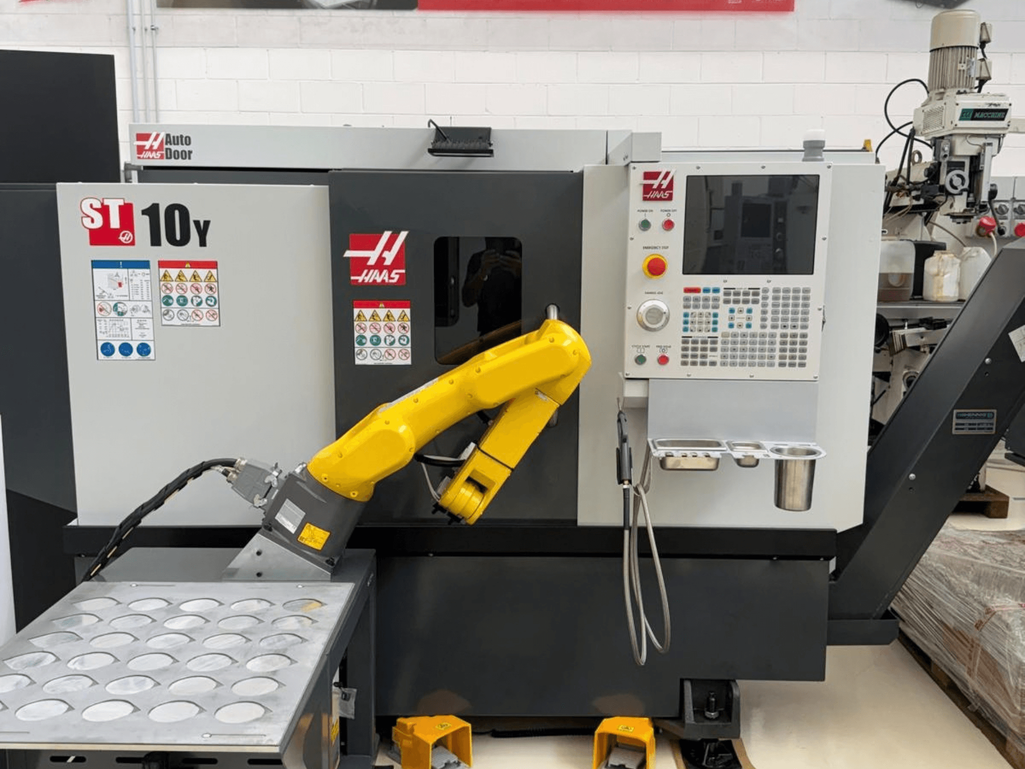 A Haas ST-10Y vízszintes eszterga elölről nézve, robotkarral és szerszámtartó tálcákkal ellátott vezérlőpanellel.