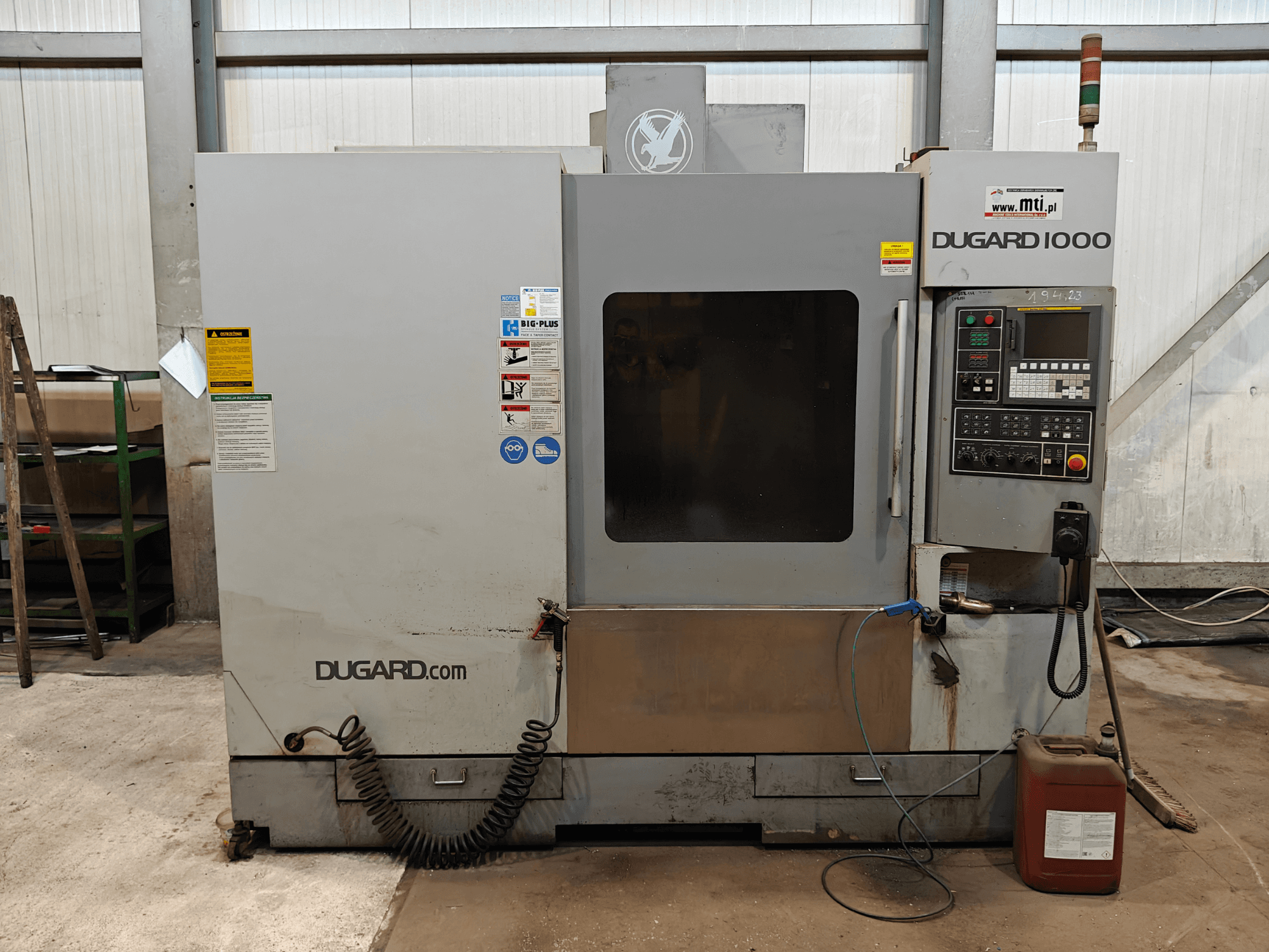 Dugard 1000 CNC gép, előlnézet, a vezérlőpanel, a biztonsági címkék és az olajtartály műhelyben.