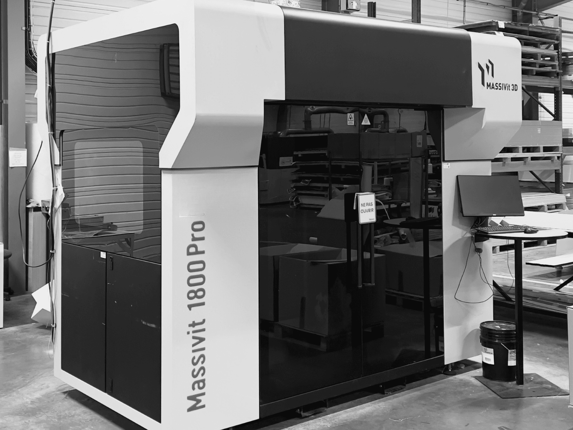 A MASSIVIT 1800 PRO műanyag 3D-nyomtató egy műhelyben, elölnézetből, látható a számítógépes munkaállomás és a vezérlőpanel.