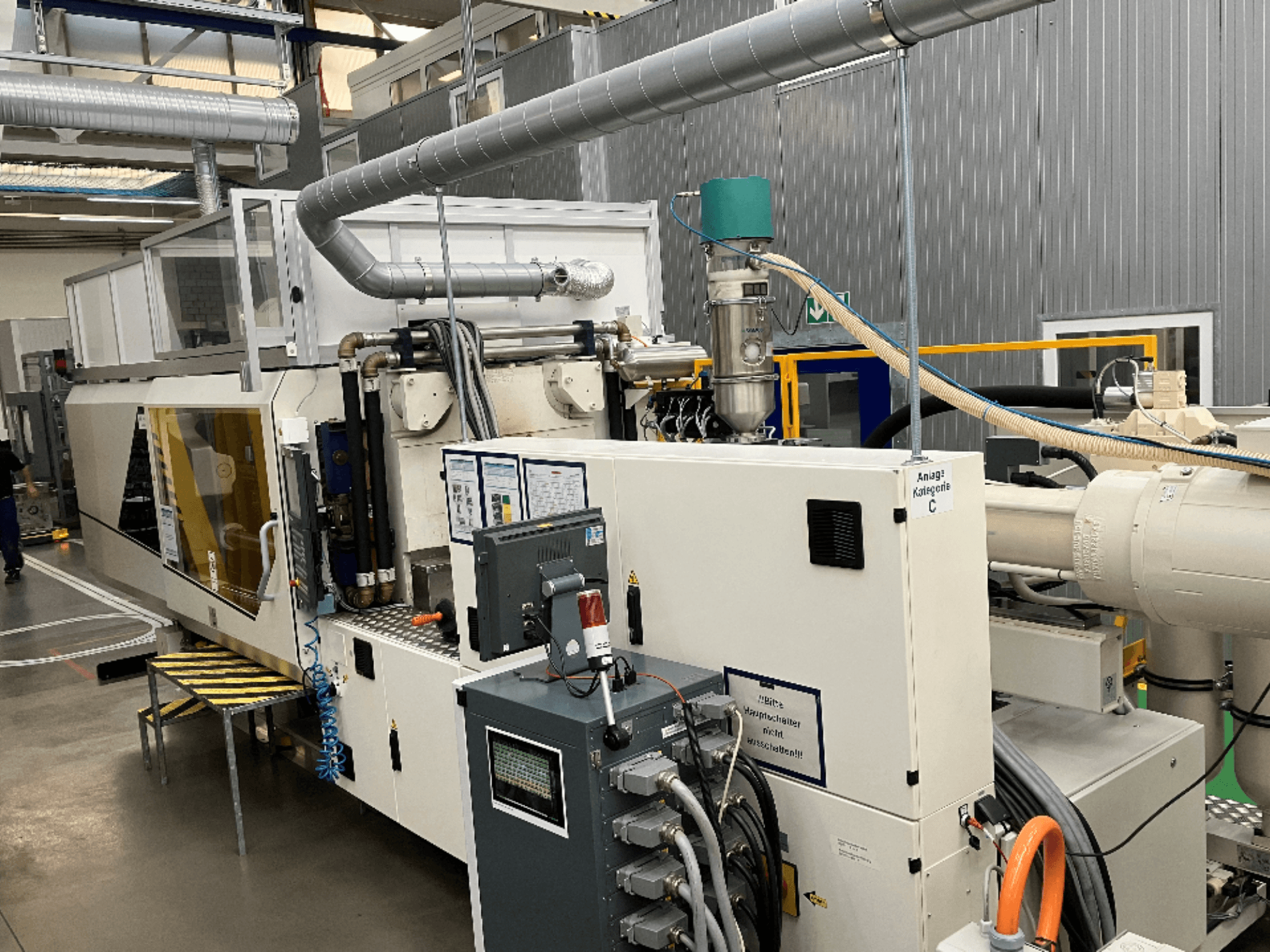 Fröccsöntőgép, elölnézetben, a márkajelzéssel, a vezérlőpanellel és a látható tömlőkkel, gyári környezetben.