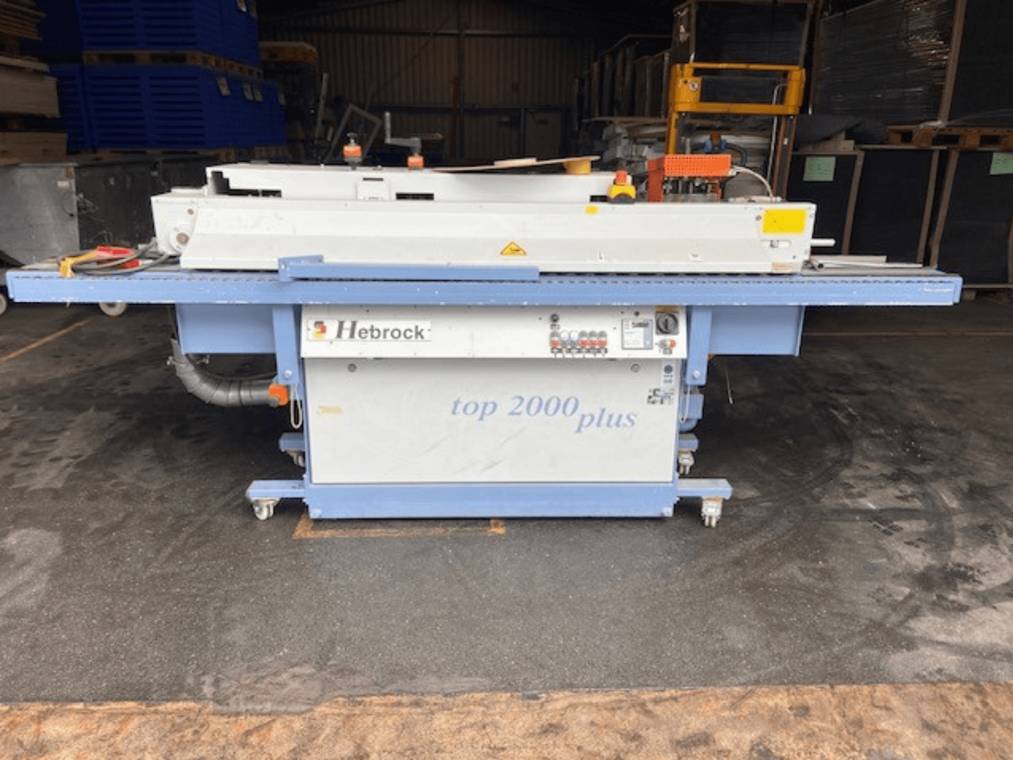 Hebrock Top 2000 Plus, elölnézet; ipari elektromos gép vezérlőpanellel, kerekekkel a mobilitás érdekében és látható biztonsági címkékkel.
