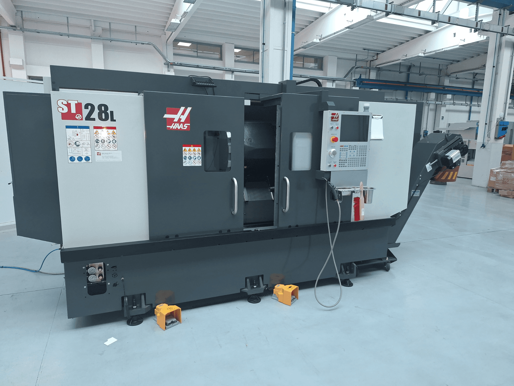 A Haas ST-28L vízszintes eszterga oldalnézete, amelyen látható a vezérlőpanel, a kezelőgombok és a biztonsági címkék.