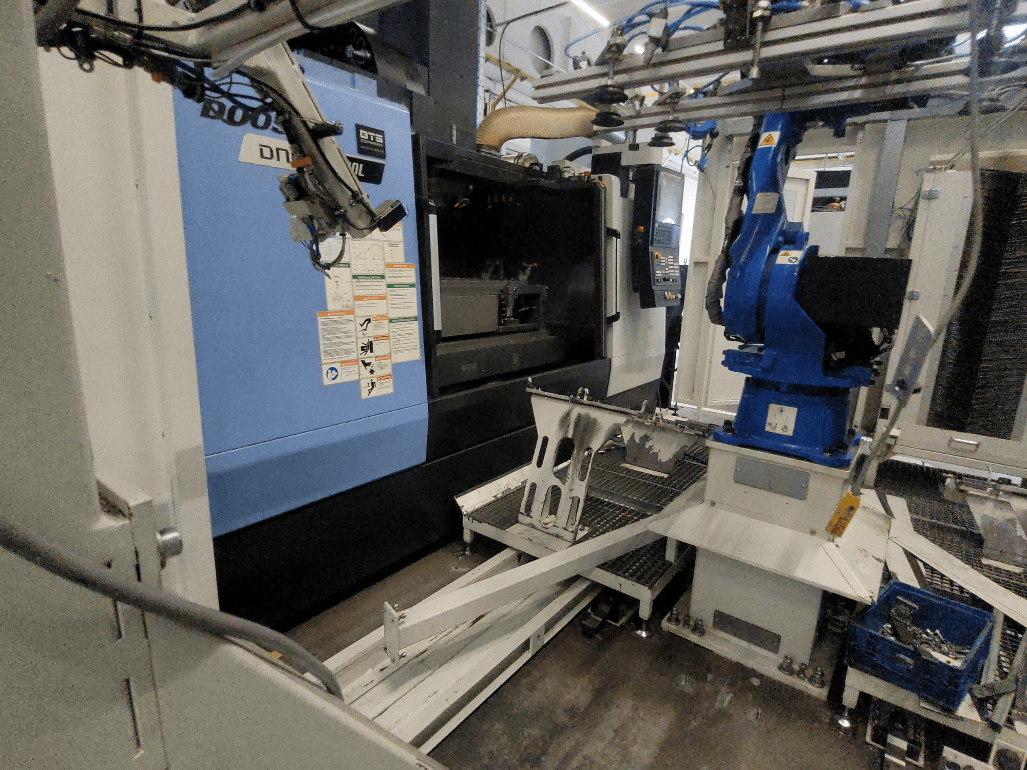 A DOOSAN DNM 6700L függőleges megmunkáló központ oldalnézete, amelyen látható a robotkar és a vezérlőpanel, bemutatva a gép felépítését.