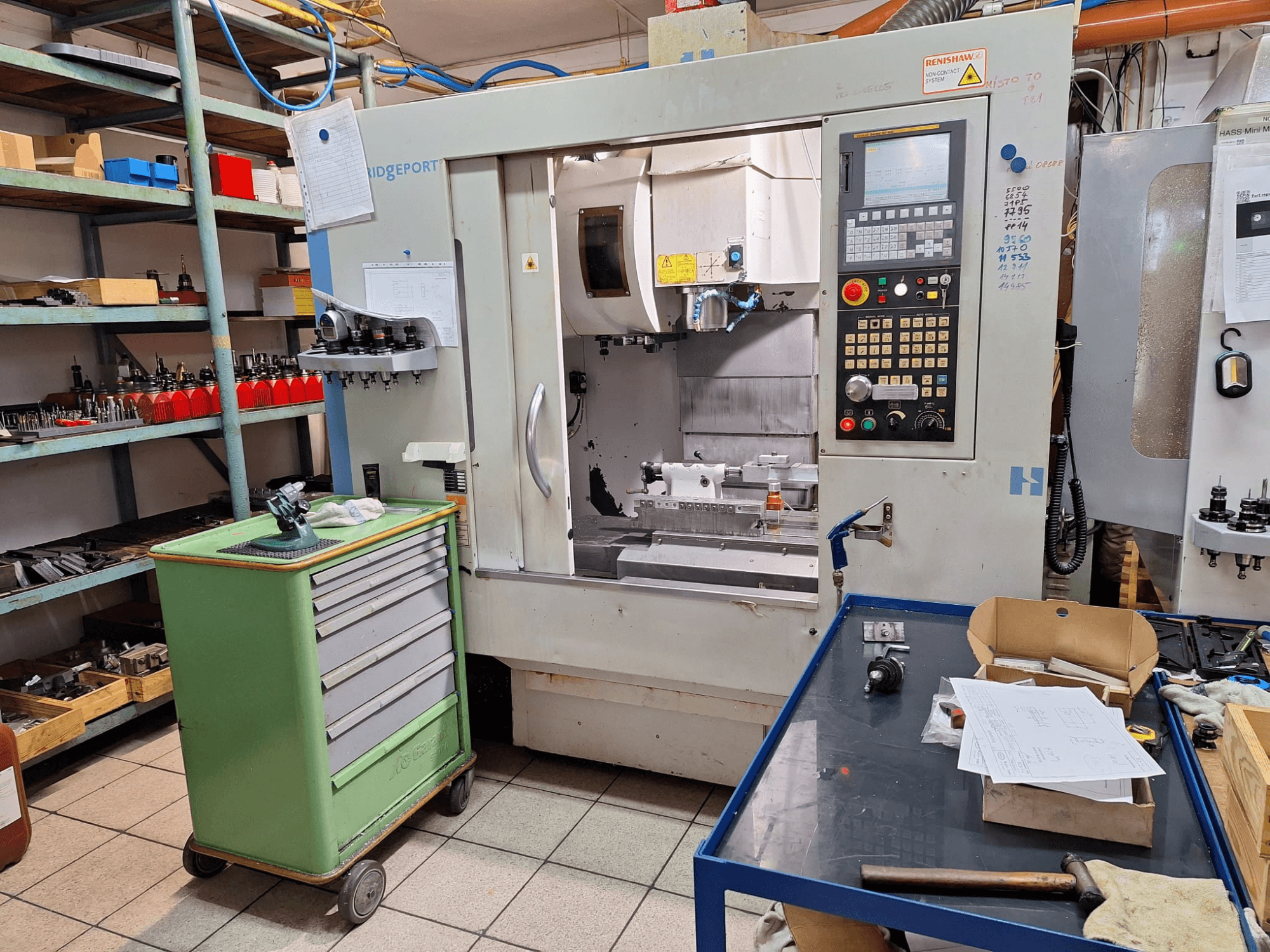 A Hardinge Bridgeport GX 710 függőleges megmunkálóközpont elölnézete vezérlőpanellel, szerszámokkal és polcokkal egy műhelyi környezetben.