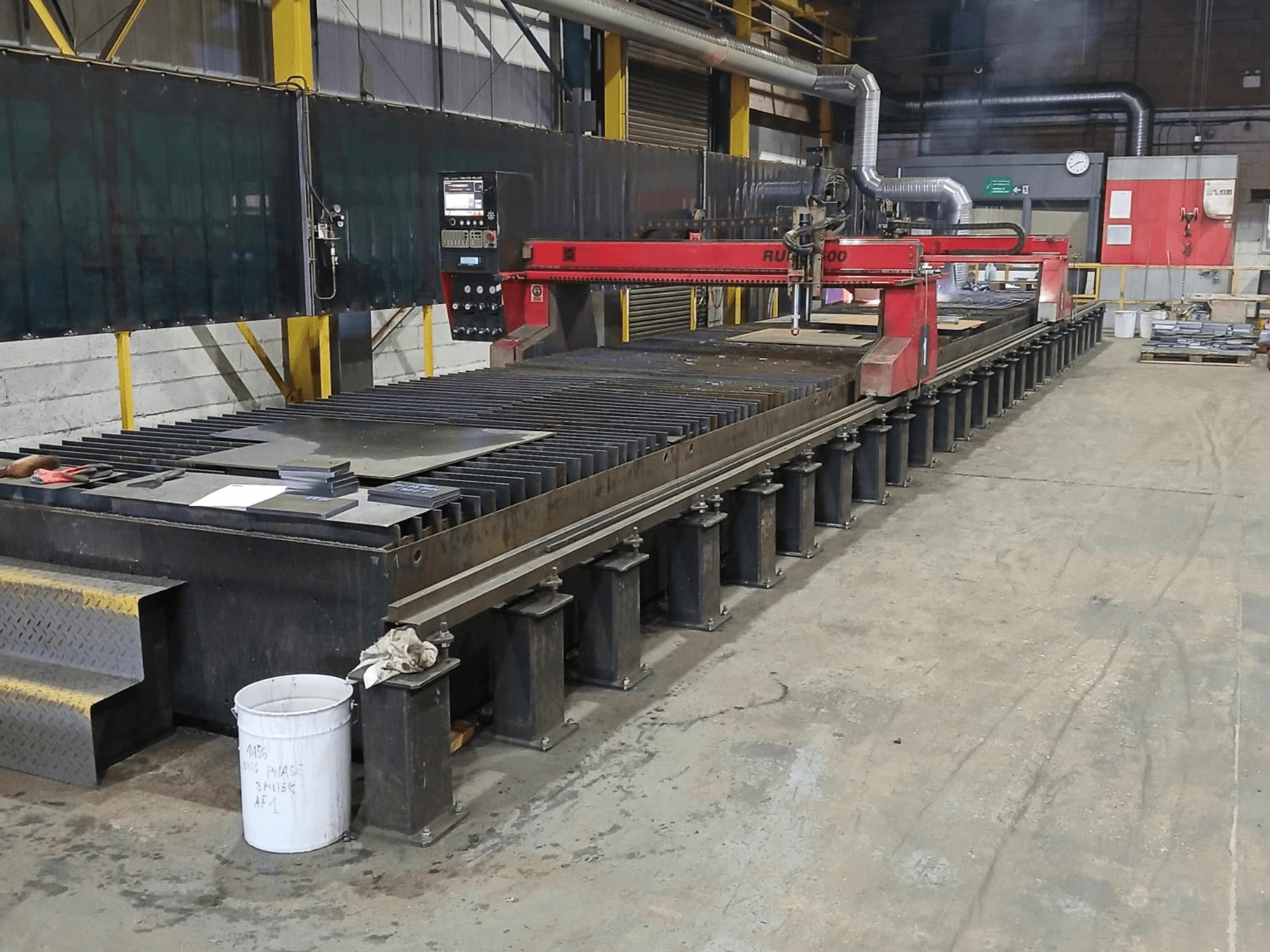 Vörös RUIDA 300 vágógép, oldalnézet, fémlemezzel egy acél asztalon, egy ipari munkaterületen.