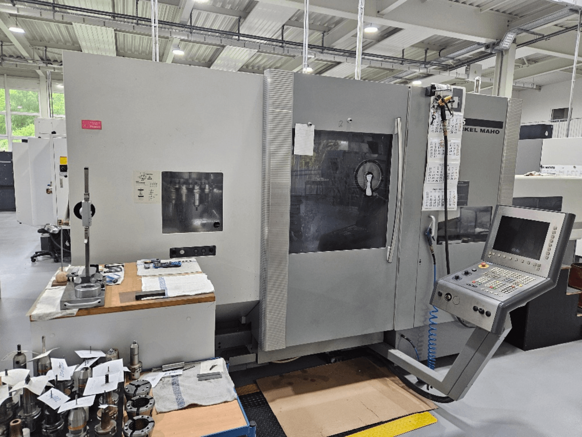 Egy DMG Mori DMU 70 gép előlnézetből, vezérlőpanellel, szerszámokkal egy faasztalon és különböző munkadarabokkal a közelben.