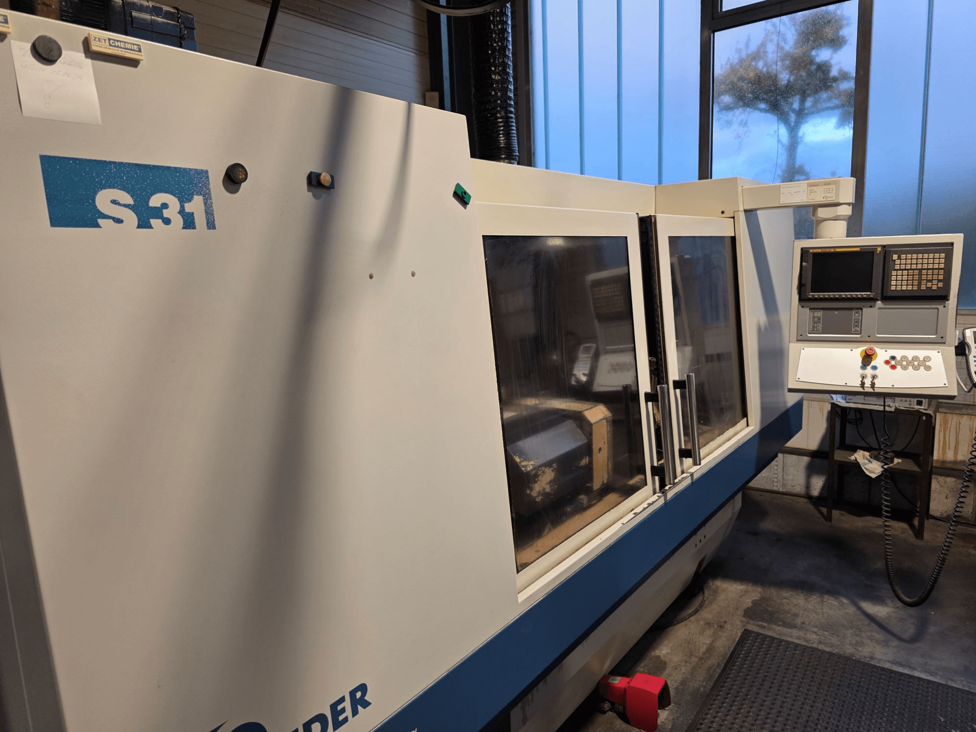 S31 CNC csiszológép, elölnézet, képernyős és gombos kezelőpanel, nagy üvegajtó, ipari környezet.