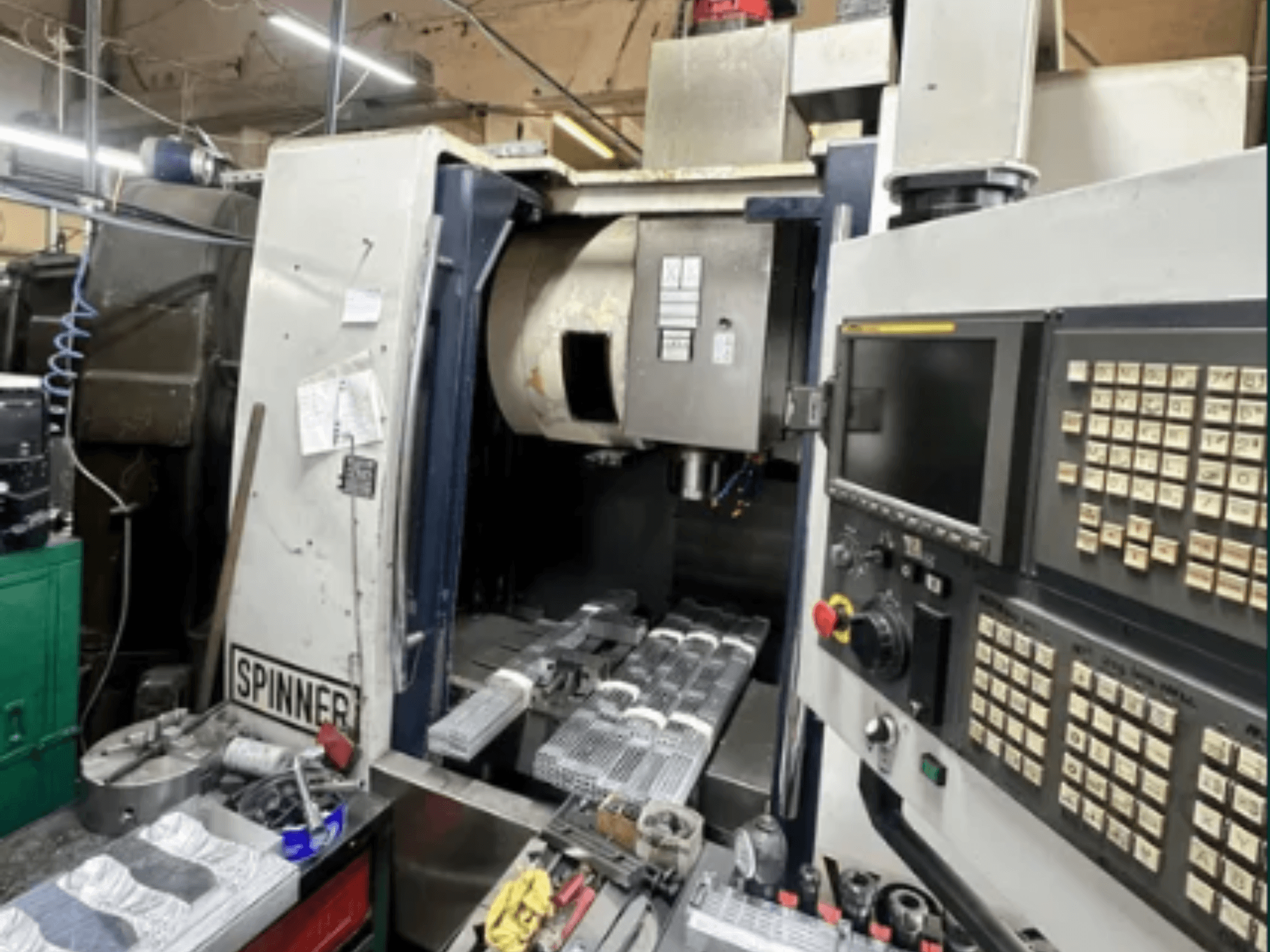 Spinner CNC-gép, előlnézet; érintőképernyős kezelőfelület, szerszámok a munkaterületen és megmunkált, feldolgozásra kész alkatrészek.