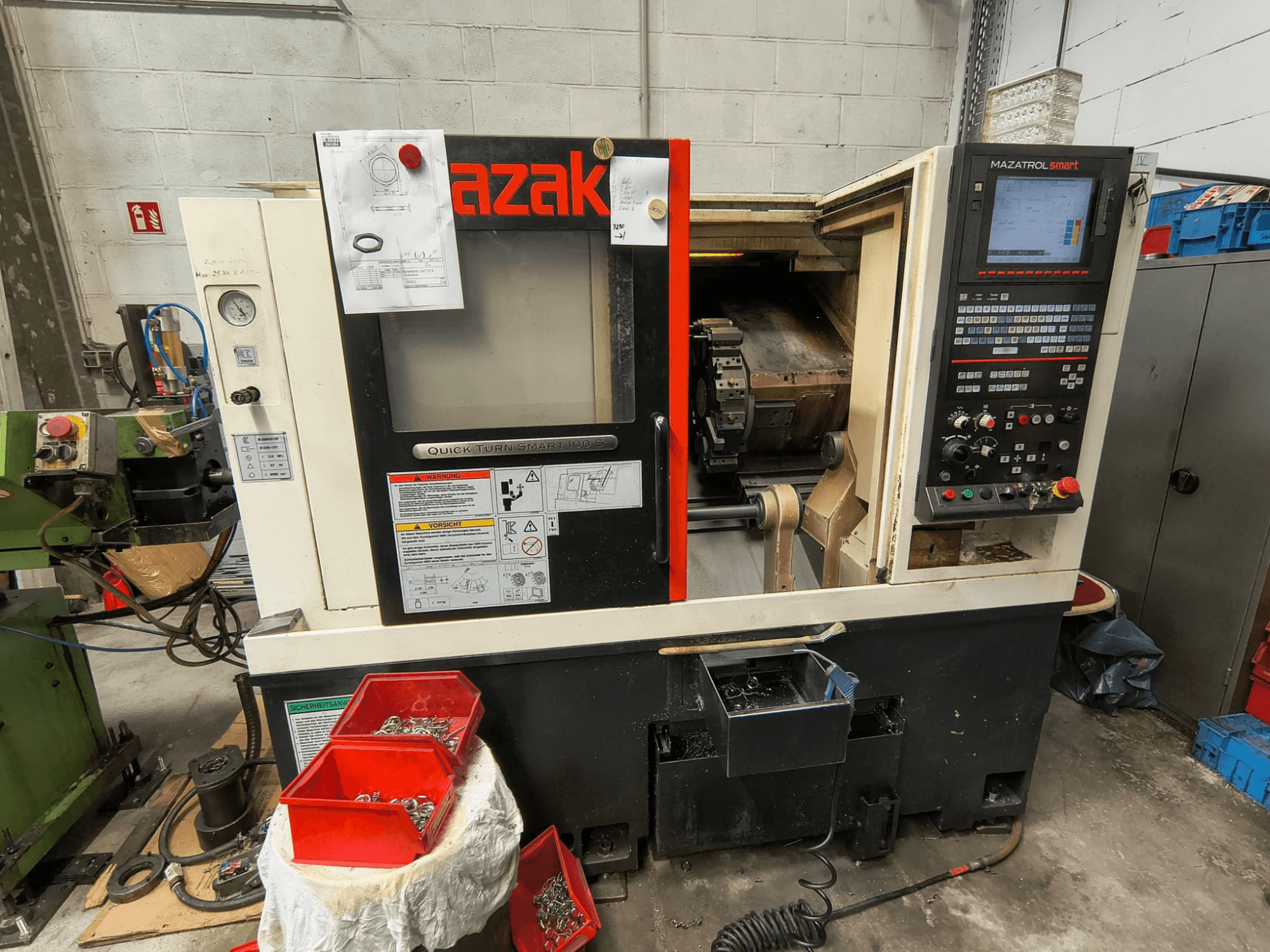 Mazak Quick Turn Smart 100 S, oldalnézet, amelyen látható a vezérlőpanel, a hűtőfolyadék-tálca és a csavarokat tartalmazó tárolórekeszek.