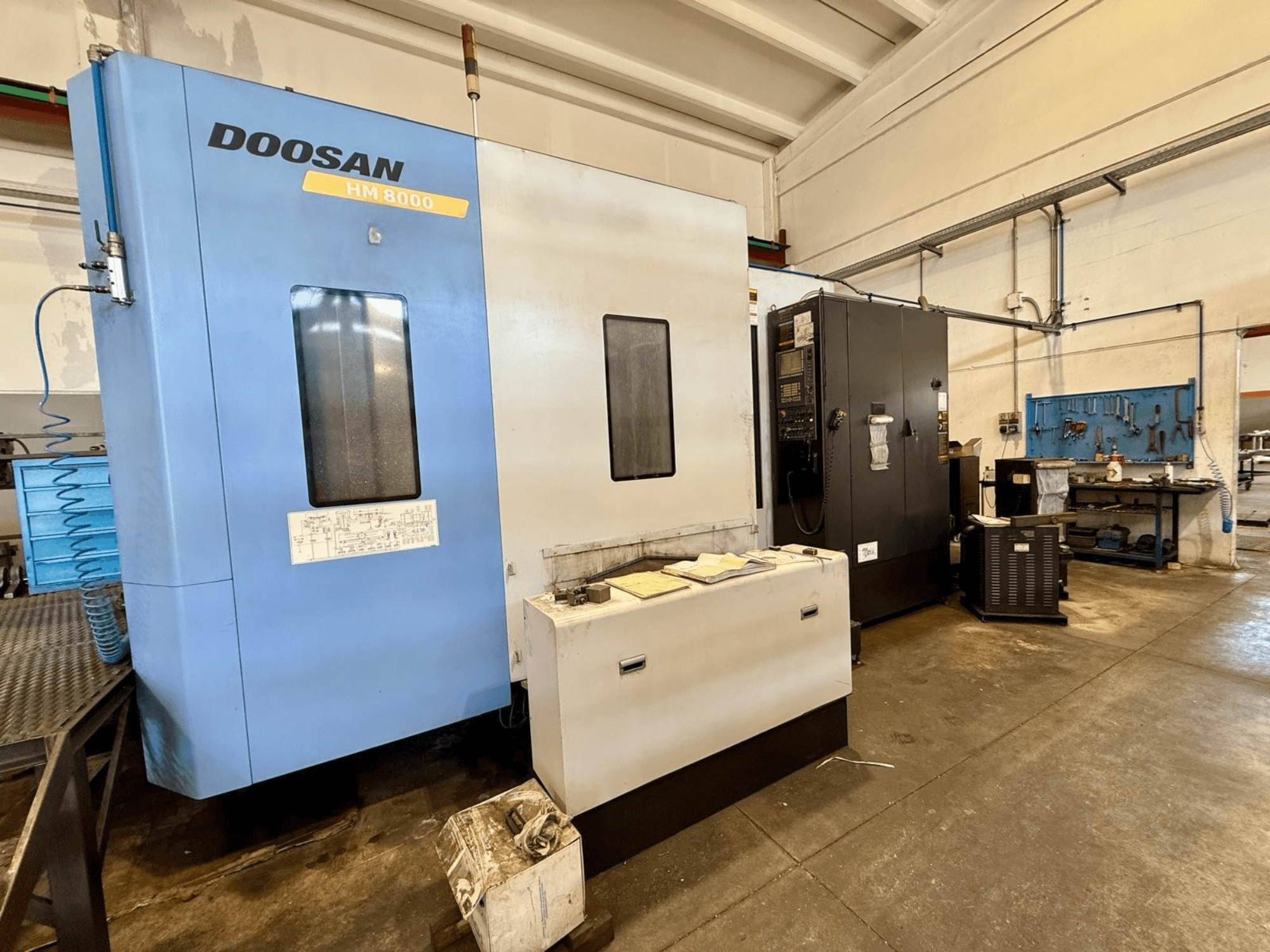 Kék színű DOOSAN HM 8000 vízszintes megmunkáló központ, elölnézetből, a vezérlőpanellel és a közelben található műhelyberendezésekkel.