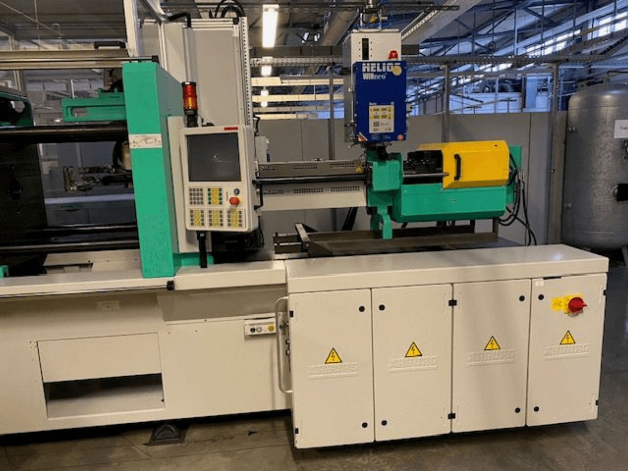 Ipari fröccsöntőgép, HELIO modell, előlnézet, vezérlőpanel, biztonsági védőburkolatok és vészleállító gomb.