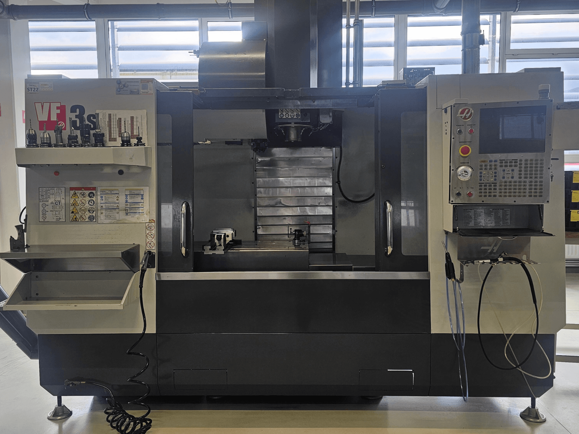 Haas VF3SS CNC-malom, előlnézet, szerszámtartókkal, vezérlőpanellel és üres hűtőfolyadék-tálcával, látható biztonsági címkékkel.