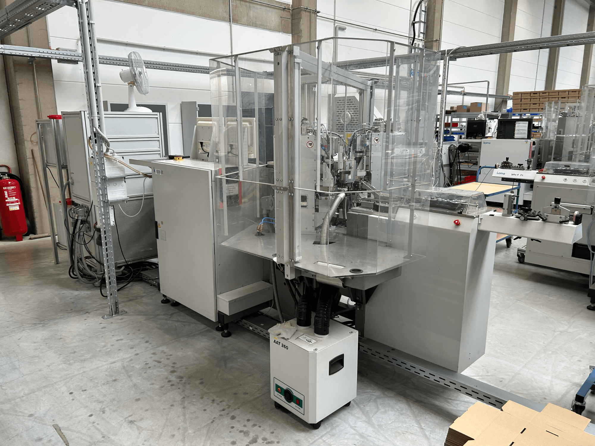 A KOMAX Alpha 433 L automatizálási berendezés gyári környezetben. Átlátszó biztonsági burkolattal és különféle vezérlőpanelekkel rendelkezik.