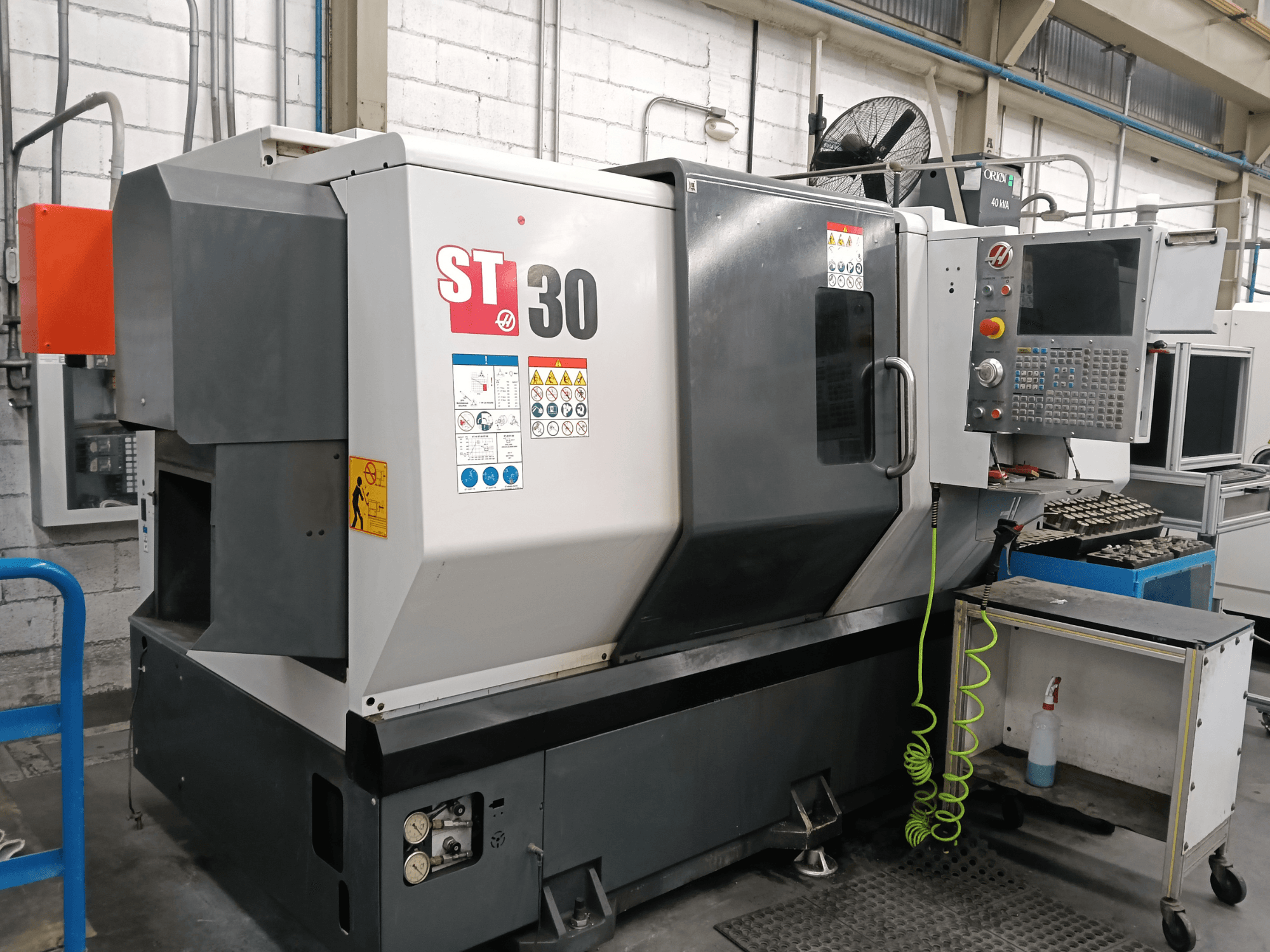Alternatív szöveg: A HAAS ST-30 vízszintes eszterga elölnézete, a vezérlőpanellel, a biztonsági címkékkel és a közeli kocsin elhelyezett szerszámokkal.