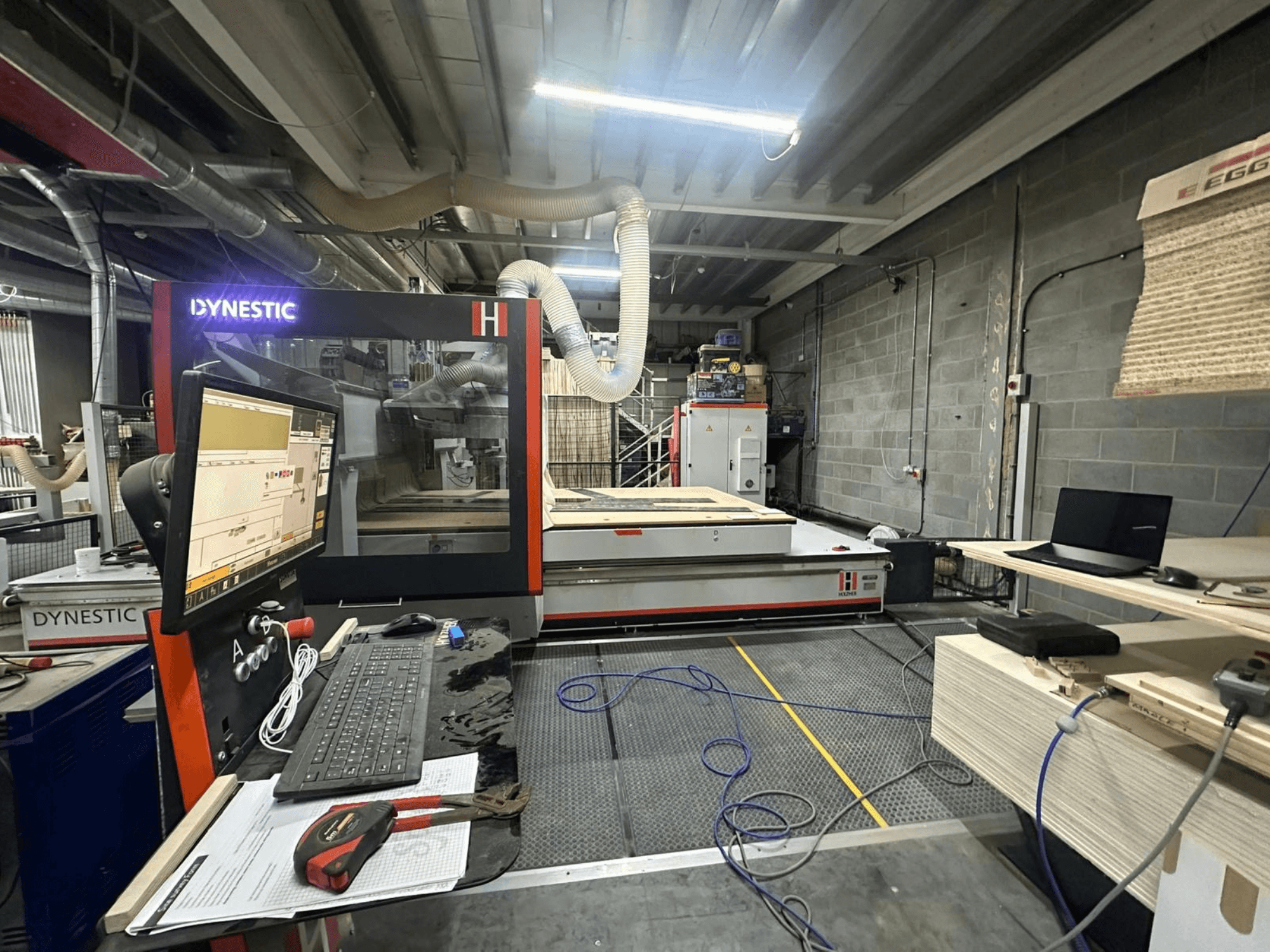 HOLZHER Dynestic 7535 fafűrészgép műhelyben, a vezérlőpanel, a monitor és az anyagok a munkaasztalon, elölnézetben.