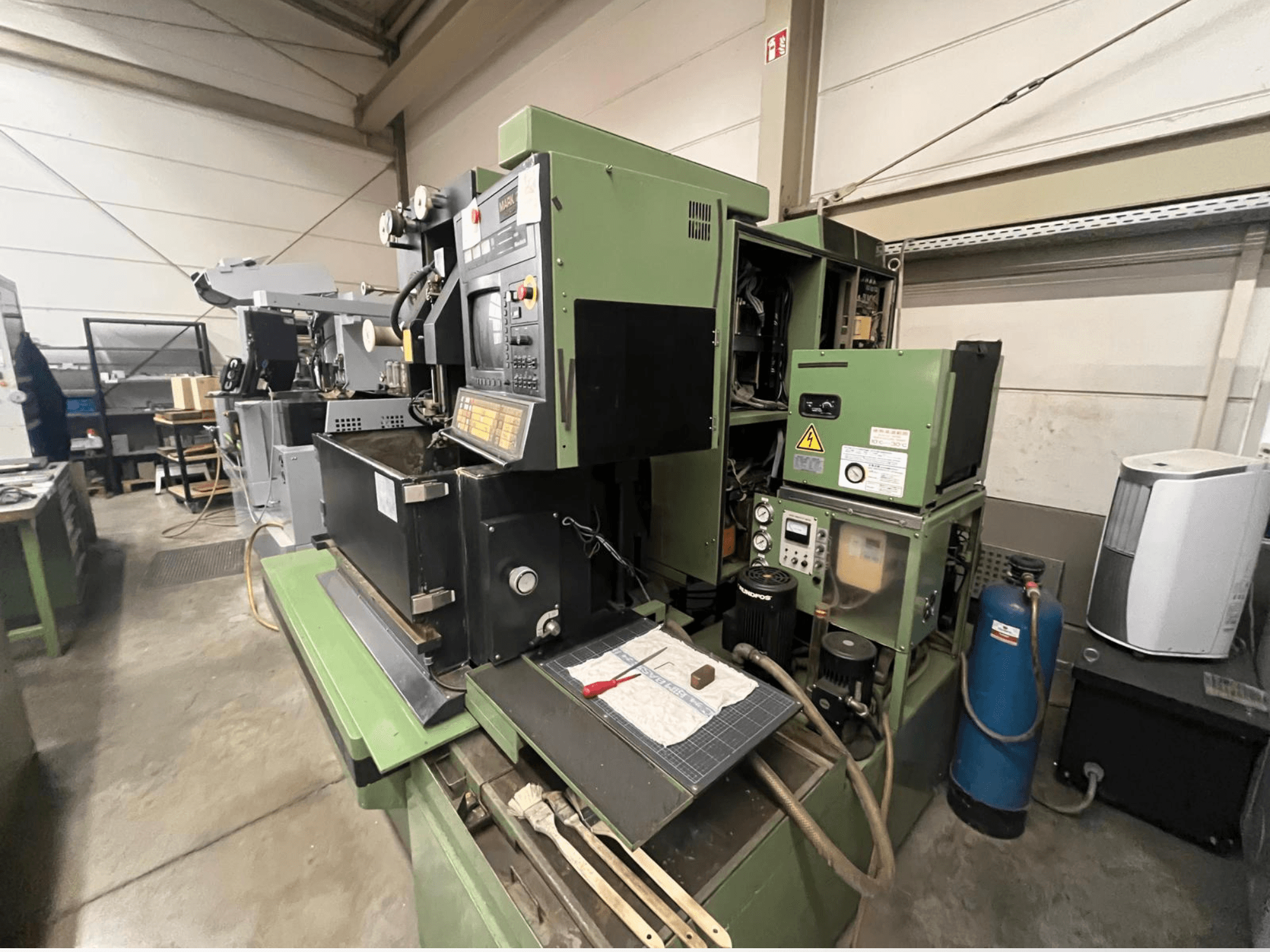 Zöld ipari gép vezérlőpanellel, oldalnézet; hidraulikus alkatrészeket és szerszámokkal ellátott munkaterületet tartalmaz.