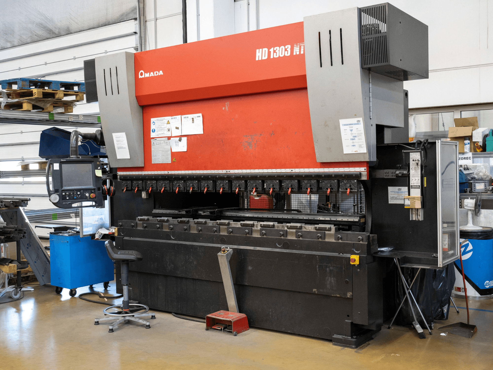 Amada HD 1303 hidraulikus présfék egy műhelyben, elölnézetben; több szorítóval, vezérlőpanellel és állítható zsámollyal rendelkezik.