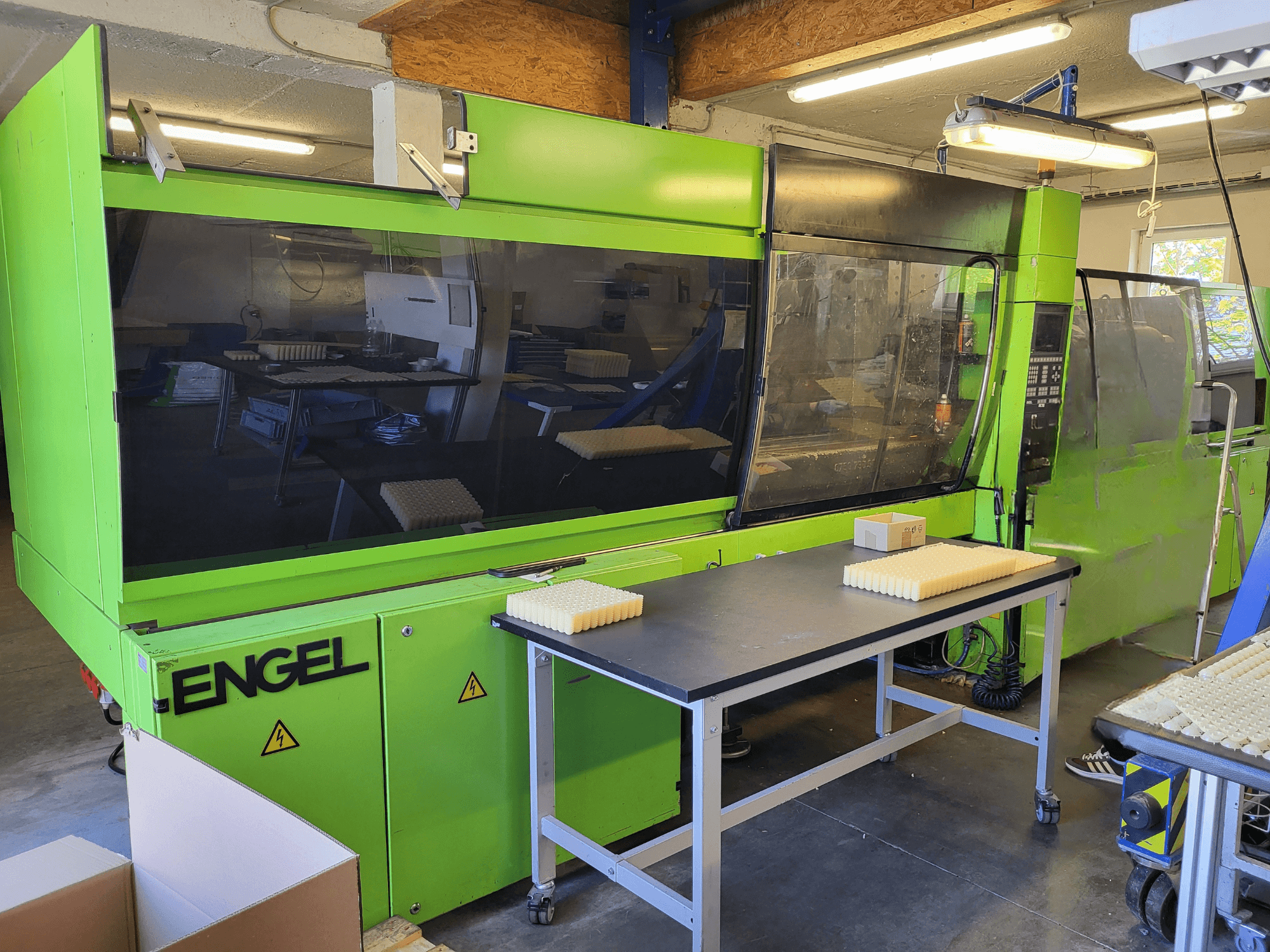 A Green Engel ES1800/300HL hidraulikus fröccsöntőgép elölről nézve, az asztalon egymásra rakott anyagokkal.