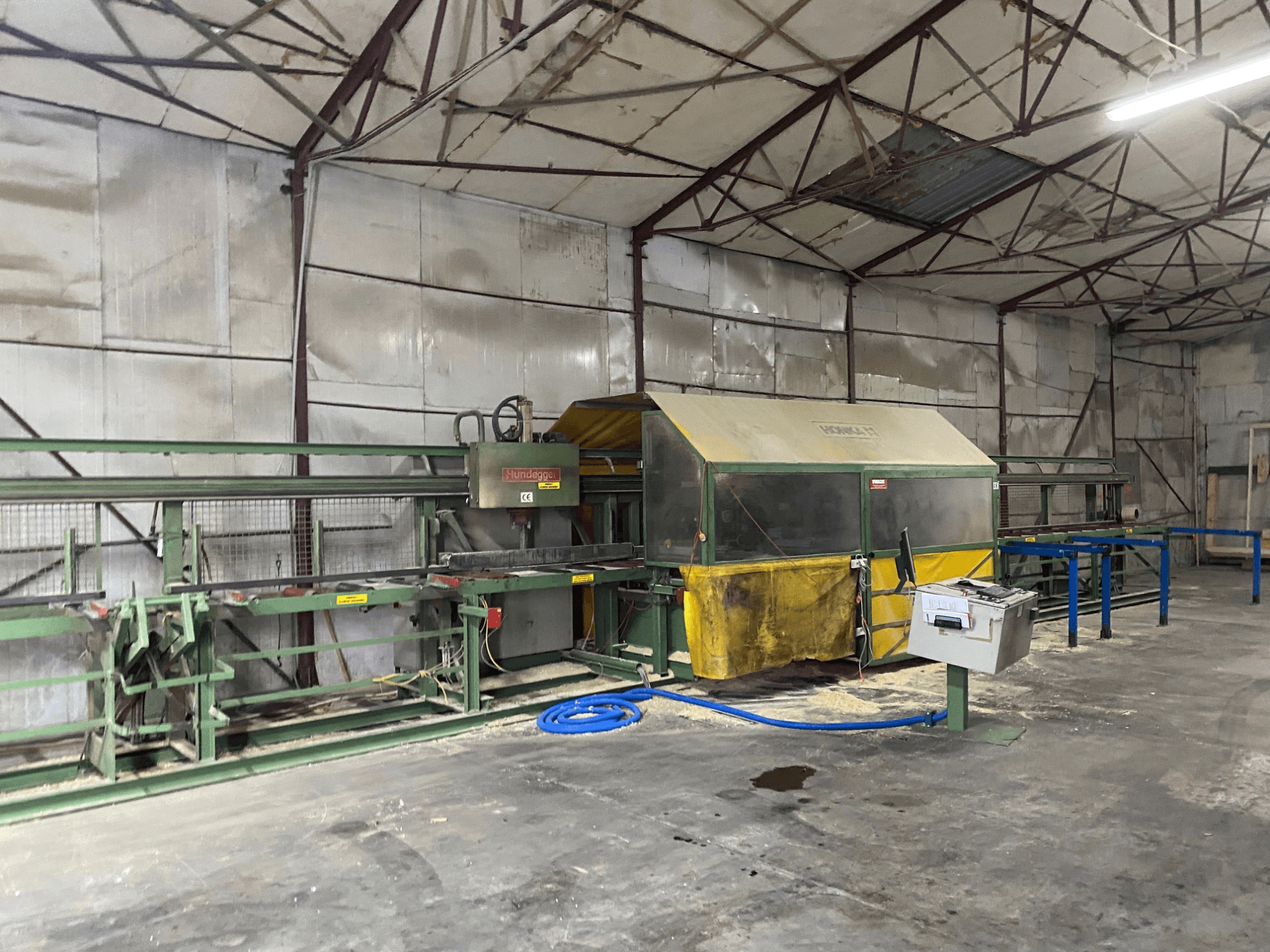 CNC faipari megmunkálóközpont HUNDEGGER K1 egy műhelyben, oldalnézetben a gépek, a kezelőszervek és a fűrészpor a padlón.