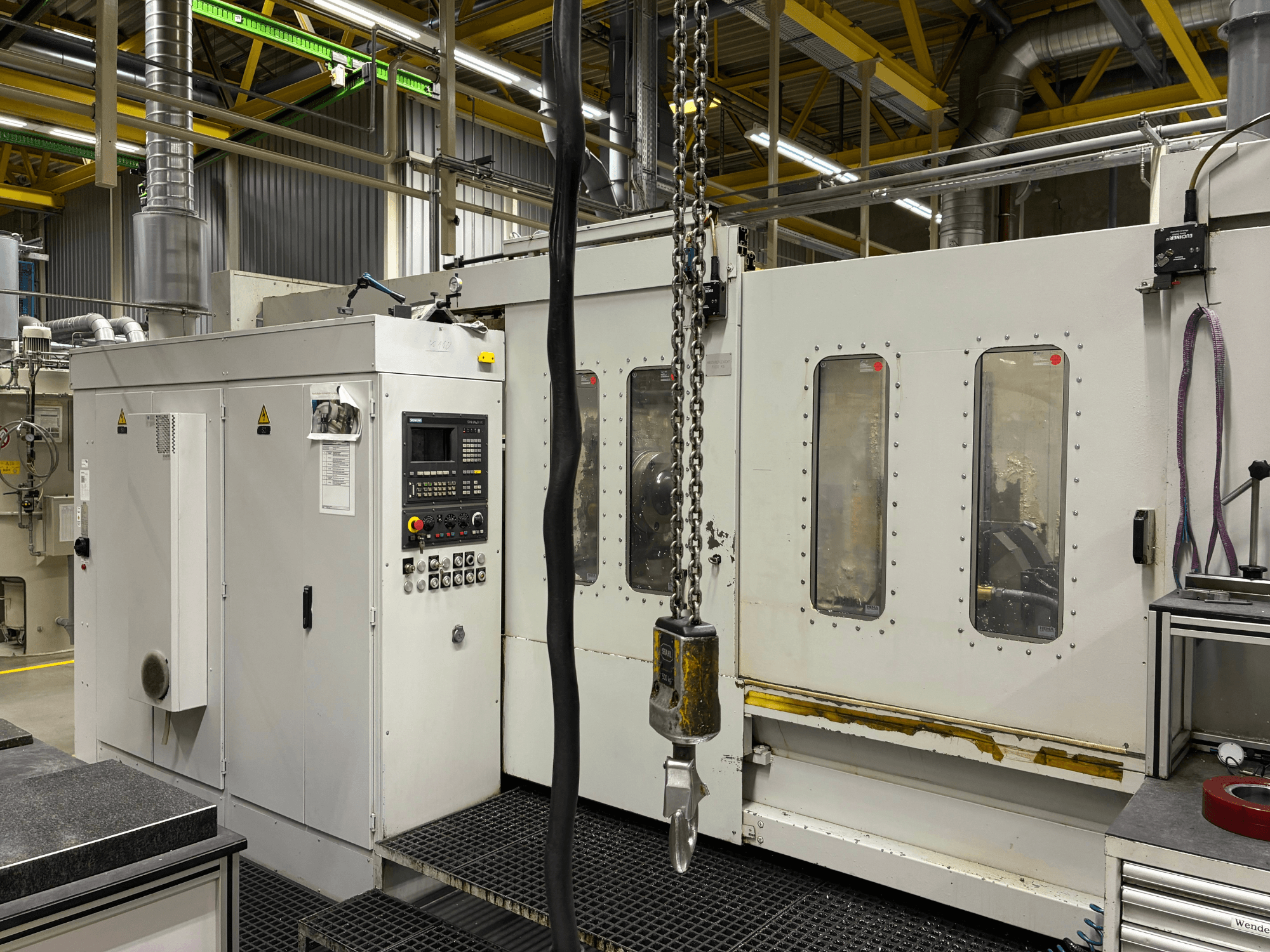 Ipari CNC-gép vezérlőpanellel, oldalnézetben; ablakok, láncok és fém alkatrészek gyári környezetben.