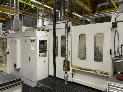 Ipari CNC-gép vezérlőpanellel, oldalnézetben; ablakok, láncok és fém alkatrészek gyári környezetben.